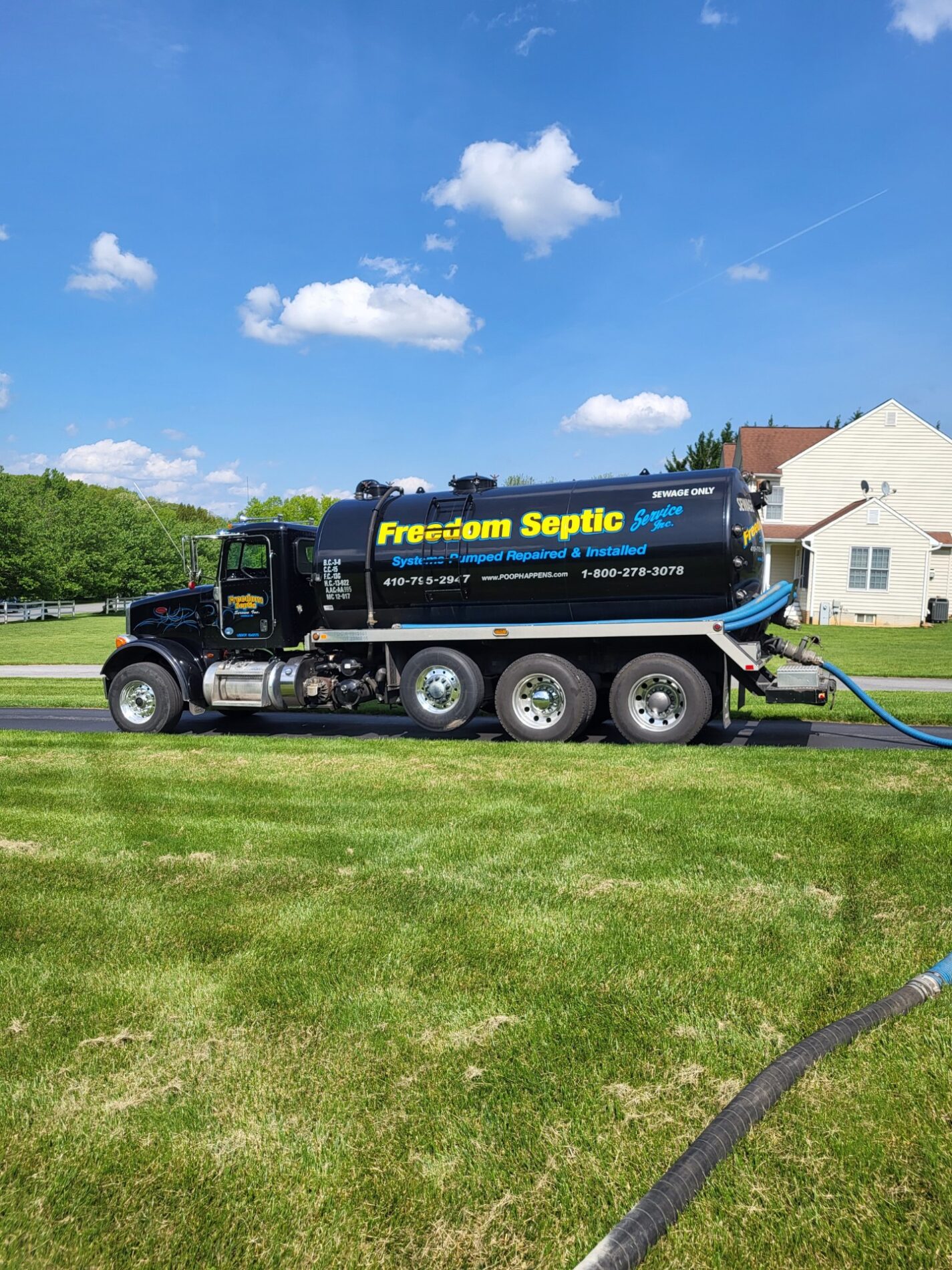 A black Freedom Septic truck parked on a grass lawn in front of a house, with a hose connected to it. The sky is clear with a few clouds.