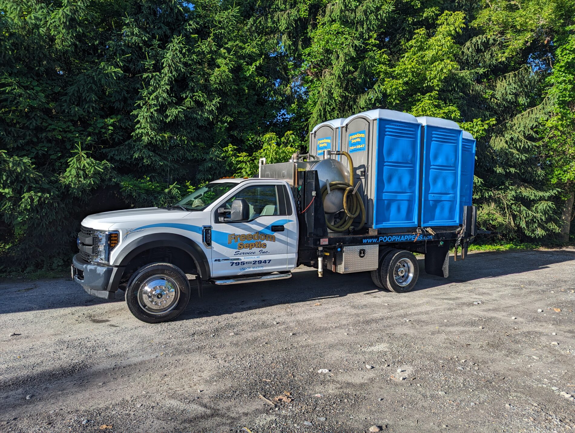 A truck with two portable toilets on the back is parked on a gravel surface in front of a wooded area. The truck has a company logo and contact information on its side.