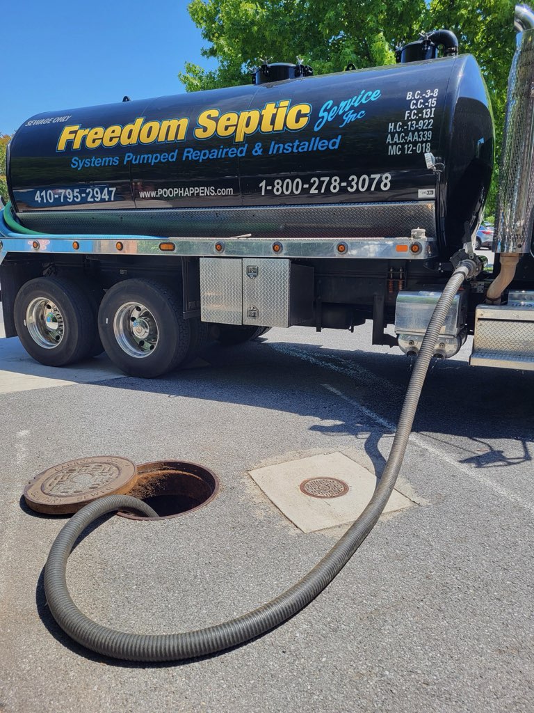 A Freedom Septic Service truck is parked near an open manhole cover with a hose inserted, apparently performing septic system maintenance. The truck has contact information and service details written on it.